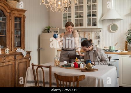 Lächelnde Frau, die einen Becher hält, mit einem Mann, der Kekse am Tisch in der Küche dekoriert Stockfoto