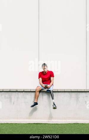 Mann, der mit einer Beinprothese sitzt und ein Smartphone an der Wand benutzt Stockfoto