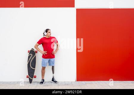 Junger Mann, der kabellose Kopfhörer trägt und mit einer Beinprothese in der Nähe des Skateboards vor der Wand steht Stockfoto