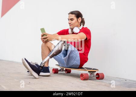 Lächelnder junger Mann, der auf Skateboard sitzt und mit einer Beinprothese sitzt und das Smartphone benutzt Stockfoto