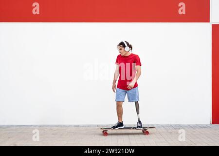 Junger Mann mit schnurlosen Kopfhörern und Skateboarden mit einer Beinprothese vor der Wand Stockfoto