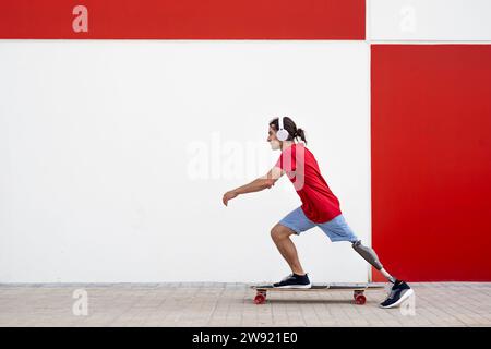 Mann mit schnurlosen Kopfhörern und Skateboarding mit einer Beinprothese vor der Wand Stockfoto