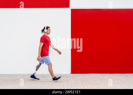 Mann mit schnurlosen Kopfhörern und Gehen mit einer Beinprothese vor der Wand Stockfoto