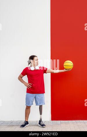 Lächelnder Mann mit einer Beinprothese, der gelben Basketball in der Nähe der Wand hält Stockfoto