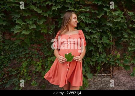 Glückliche schwangere Frau, die ein pfirsichfarbenes Kleid trägt und Ahornblatt vor Pflanzen hält Stockfoto