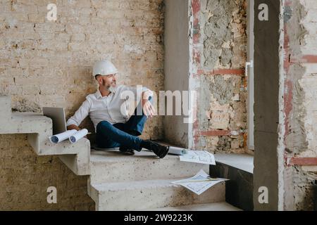 Lächelnder Architekt sitzt mit Laptop und Bauplänen auf Treppen vor Ort Stockfoto