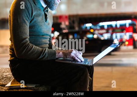 Freiberufler mit Laptop, der nachts in der Stadt auf der Bank sitzt Stockfoto