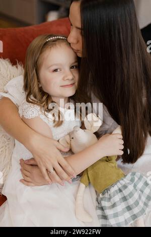 Mutter küsst lächelnde Tochter, die zu Hause mit Kuschelspielzeug sitzt Stockfoto
