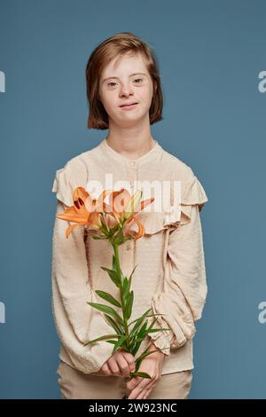 Teenager-Mädchen mit Down-Syndrom, das orangene Orchideenblüten vor blauem Hintergrund hält Stockfoto