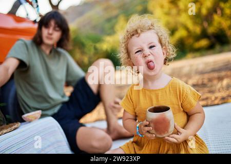 Niedliches Mädchen mit Teetasse, die die Zunge ausragt, bei Bruder sitzend Stockfoto