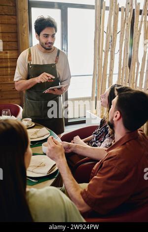 Junger Kellner mit Tablet-PC, der Bestellungen von Gästen im Restaurant entgegennimmt Stockfoto