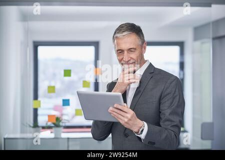 Lächelnder Geschäftsmann mit Hand am Kinn mit Tablet-PC im Büro Stockfoto