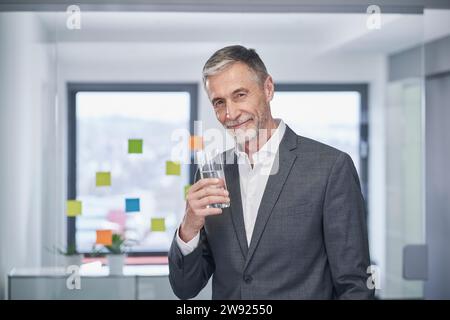 Lächelnder Geschäftsmann, der mit einem Glas Wasser im Büro steht Stockfoto