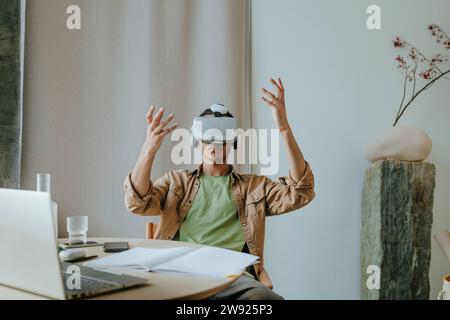 Junger Mann mit VR-Headset, der mit erhobenen Armen am Tisch sitzt Stockfoto