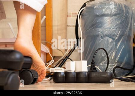Ein Überspannungsschutz liegt auf dem Boden in der Wohnung und verschiedene Stecker von elektronischen Geräten sind eingeklemmt, weibliche Beine in der Nähe, Kabel und eine Ladung Stockfoto