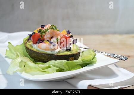 Gefüllte Avocado mit Garnelen, Tomaten, Mayonnaise und Kresse garniert auf Salat und einem weißen Teller, heller rustikaler Hintergrund, festliche Vorspeise oder Holida Stockfoto
