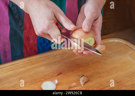 Hände einer Frau, die eine Zwiebel mit einem Küchenmesser über einem hölzernen Schneidebrett schält, Vorbereitung zum Kochen zu Hause, Kopierraum, ausgewählter Fokus, Erzähler Stockfoto