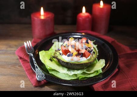 Halbe Avocado gefüllt mit Garnelen, Tomaten und Mayonnaise auf einem schwarzen Teller, rote Kerzen und Serviette, dunkler rustikaler Holzhintergrund, festlicher Feiertag A Stockfoto