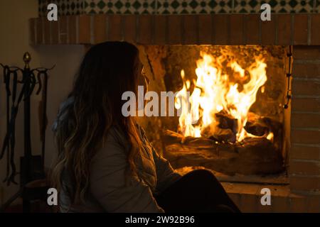 Lateinische Frau, die in den kalten Wintern vor dem Kamin sitzt, in den Flammen vertieft Stockfoto