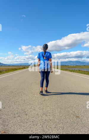 Ein Reisender in lässiger Kleidung, der auf einer asphaltierten Straße in Richtung Horizont in einer ruhigen Umgebung spaziert, Rückblick auf Latina-Frau, die in blau gekleidet ist Stockfoto