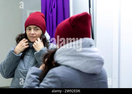 Frau mittleren Alters, die in einem Umkleideraum an einer Abholstelle steht, eine stylische Jacke und einen Hut anprobiert und in den Spiegel blickt, um die Passform und zu beurteilen Stockfoto