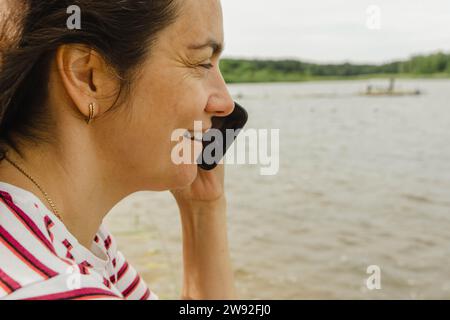 Porträt einer Frau mittleren Alters, die auf einem Handy spricht, während sie sich am See entspannt Stockfoto