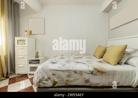 Ein Schlafzimmer mit einem Einzelbett mit bedruckten Federdecken, mehreren Kissen, weißen Möbeln, Klappcouch, Steinzeugfußboden und Vorhängen an den Fenstern Stockfoto