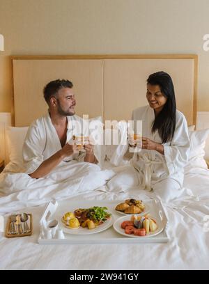 Paare, die im Bett eines Luxushotels frühstücken, Männer und Frauen im Bett mit Frühstückskaffee und Brot. Ein vielfältiges Paar einer asiatischen Frau und eines europäischen Mannes im Schlafzimmer Stockfoto