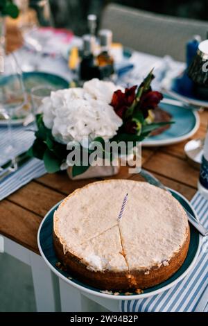 Kuchen mit einem ausgeschnittenen Sektor und einer Kerze steht auf einem Teller neben einem Blumenstrauß auf dem Tisch Stockfoto