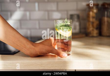 Nahaufnahme der weiblichen Hand hält einen erfrischenden Zitronen-Mojito auf einem Holztisch in einer Küche zu Hause. Eis, Minze und Limette garnieren den sommerlichen Cocktail. Stockfoto