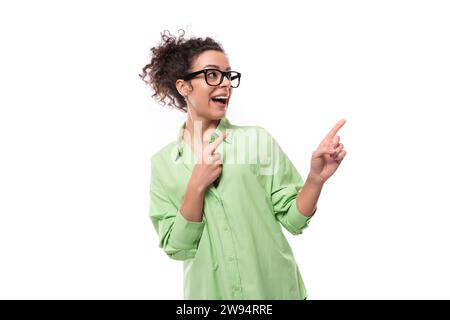 Hübsche junge, brünette lockige Frau in Brille in einem grünen Hemd zeigt ihre Hand zur Seite auf einem weißen Hintergrund mit Kopierraum Stockfoto