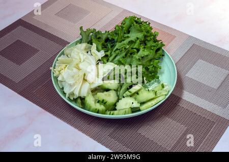 Frischer Sommersalat mit Frühkohl, Gurken und Salat auf grünem Teller auf braunem Tischset. Grüner Gemüsesalat auf einem Teller. Gesunde Ballaststoffe Stockfoto