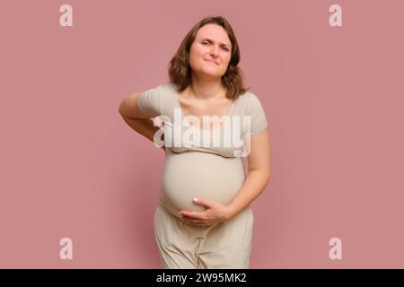 Eine schwangere Frau hält ihre Schmerzen vor einem rosa Hintergrund des Studios zurück. Schwangerschaft bei einer Frau mit Bauch Stockfoto