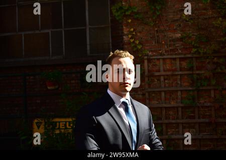 Junger muskulöser weißer Mann in einem schwarzen Anzug, weißem Hemd mit Kragen und hellblauer Krawatte, der vor einer Ziegelwand im dunklen Hintergrund posiert Stockfoto