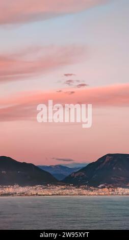 Rosa Wolken am Abendhimmel über blauem Meer auf dem Hintergrund der Stadt und Berge am Horizont, vertikaler Rahmen. Stockfoto