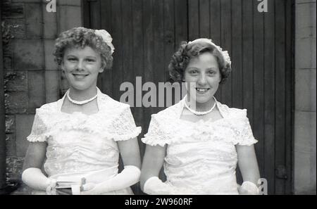 1950er Jahre, hhistorisch, Hochzeit, zwei Brautjungfern, junge Mädchen im Teenageralter, in ihren Outfits stehen sie für ein Foto an der Holztür des Kircheneingangs, England, Großbritannien. Stockfoto