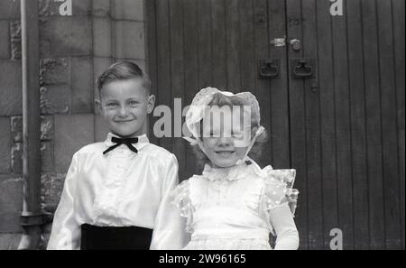 1950er Jahre, historisch, Hochzeit, großes Lächeln von der Seite Junge und Blumenmädchen, während sie zusammen stehen und Hände halten vor den Türen der Kirche, England, Großbritannien. Bei einer Hochzeitszeremonie nahm ein Pageboy an einem Brautzug Teil und trug einen Brautzug, während die traditionelle Rolle eines Blumenmädchens darin besteht, Blumen zu tragen und sie zu streuen, bevor die Braut den Gang hinunter geht. Stockfoto