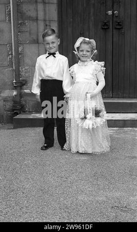 1950er Jahre, historisch, Hochzeit, großes Lächeln von der Seite Junge und Blumenmädchen, während sie zusammen stehen und Hände halten vor den Türen der Kirche, England, Großbritannien. Bei einer Hochzeitszeremonie nahm ein Pageboy an einem Brautzug Teil und trug einen Brautzug, während die traditionelle Rolle eines Blumenmädchens darin besteht, Blumen zu tragen und zu streuen, bevor die Braut den Gang hinunter geht. Stockfoto