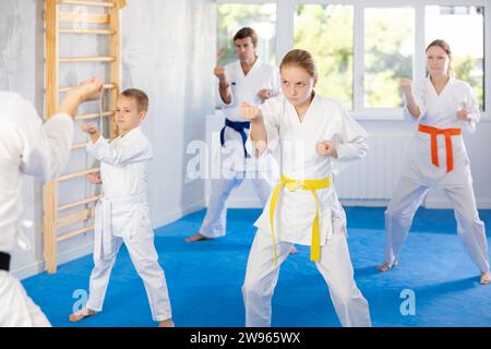 Teenager-Mädchen in weißem Kimono gekleidet mit Gürtel-Übungstechniken mit Trainer, im Hintergrund macht ihre Familie Kata Stockfoto