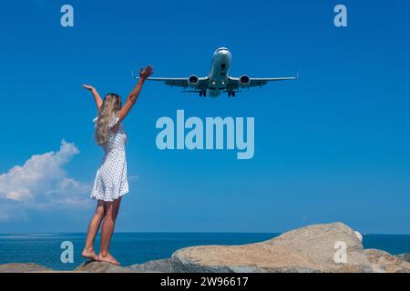 Aerial Rendezvous: Mädchen in weißem Kleid auf Steinen, Flugzeug über dem Blauen Meer Stockfoto