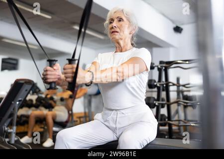 Aktive Athletin reife Frau bei Übungen am Crossover-Trainingsgerät mit Loops und Gewichtung. Trizeps, Bizeps, Deltamuskel, Brustmuskeltraining Stockfoto