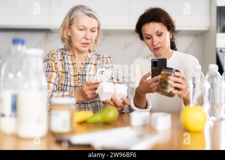 Zwei sorgfältige Frauen scannen verschiedene Produkte mit ihren Smartphones in der Küche Stockfoto