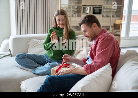 Ein überraschter Mann packt ein Geschenk aus, eine Frau wartet auf der Couch in der Nähe, mit betenden Händen, die Reaktion sehen Stockfoto
