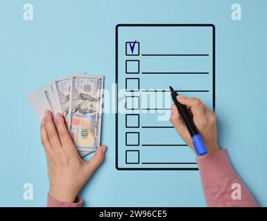 Die Hand einer Frau, die einen Stapel amerikanischer Dollar hält und eine Liste mit Einkäufen oder Ausgaben ausfüllt, um die Finanzkontrolle zu betonen. Stockfoto
