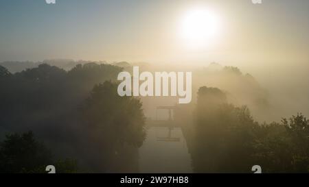 Dieses eindrucksvolle Bild fängt das zarte Gleichgewicht von Licht und Nebel bei Sonnenaufgang über einem ruhigen Fluss ein. Der Fluss selbst ist kaum zu sehen, da eine dicke Nebeldecke die Landschaft sanft umhüllt und nur die Silhouetten von Bäumen und einen Hinweis auf den Wasserweg sichtbar macht. Die Sonne, die durch den Nebel gestreut wird, sorgt für eine sanfte Hintergrundbeleuchtung, die die ruhige und mystische Qualität des frühen morgens verstärkt. Dawn ist sanfte Umarmung, nebeliger Morgen über dem Fluss. Hochwertige Fotos Stockfoto