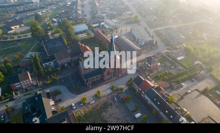 Das Bild zeigt die Sint Jozef Kirche in Rijkevorsel aus der Luft bei Sonnenaufgang. Die aufgehende Sonne wirft ein warmes Licht auf die Backsteinarchitektur der Kirche und beleuchtet das Gebäude und seinen markanten Turm. Die umliegende Wohngegend sonnt sich auch im Morgenglühen, und die Straßen warten ruhig auf die Aktivitäten des Tages. Diese Szene stellt die zeitlose Eleganz der historischen Architektur dem täglichen Leben der Stadt gegenüber. Aerial Dawn Über Sint Jozef Church, Rijkevorsel. Hochwertige Fotos Stockfoto