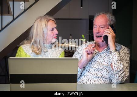 Dieses Bild zeigt ein Senior-Paar, das gemeinsam an der Lösung eines Problems arbeitet, wobei die Frau besorgt oder verwirrt aussieht, während der Mann am Telefon ist und möglicherweise mit dem Kundensupport spricht. Der Mann gibt Gesten, als würde er die Situation erklären, während die Frau um Sicherheit oder Klärung zu bitten scheint. Ihre Zusammenarbeit vor dem Hintergrund einer modernen Küche unterstreicht die Schnittstelle älterer Generationen mit moderner Technologie und Unterstützungssystemen. Senior Tech Collaboration, Problemlösung in Aktion. Hochwertige Fotos Stockfoto