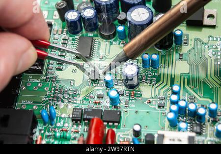 Nahaufnahme der Hände eines Technikers in einer Werkstatt. Der Servicebetrieb lötet die Leiterplatte eines elektronischen Geräts mit einer Pinzette auf dem Tisch. Stockfoto