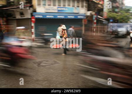 Eine Tomatenverkäuferin spaziert durch die Straßen von Hanoi, Vietnam, während Motorroller vorbeifahren. (Bewegungsunschärfe) Stockfoto