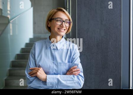 Eine lächelnde und erfolgreiche junge Geschäftsfrau steht auf dem Flur eines Bürozentrums mit überkreuzten Armen auf der Brust und lächelt, während sie zur Seite blickt. Stockfoto